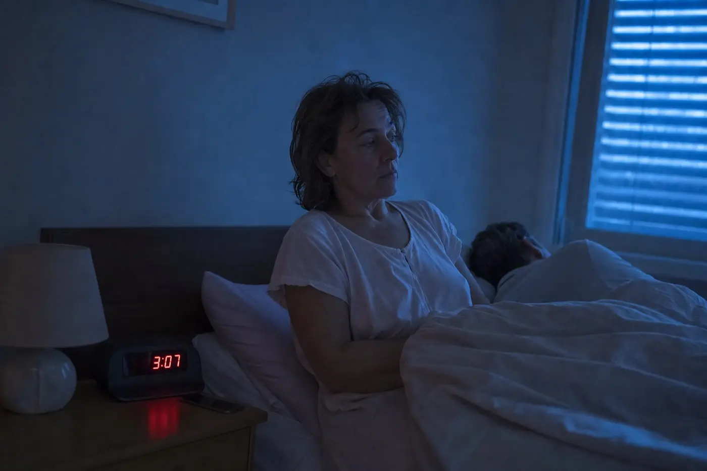 Eine Frau sitzt nachts aufrecht im Bett und schaut auf den Wecker, der 3:07 zeigt.
