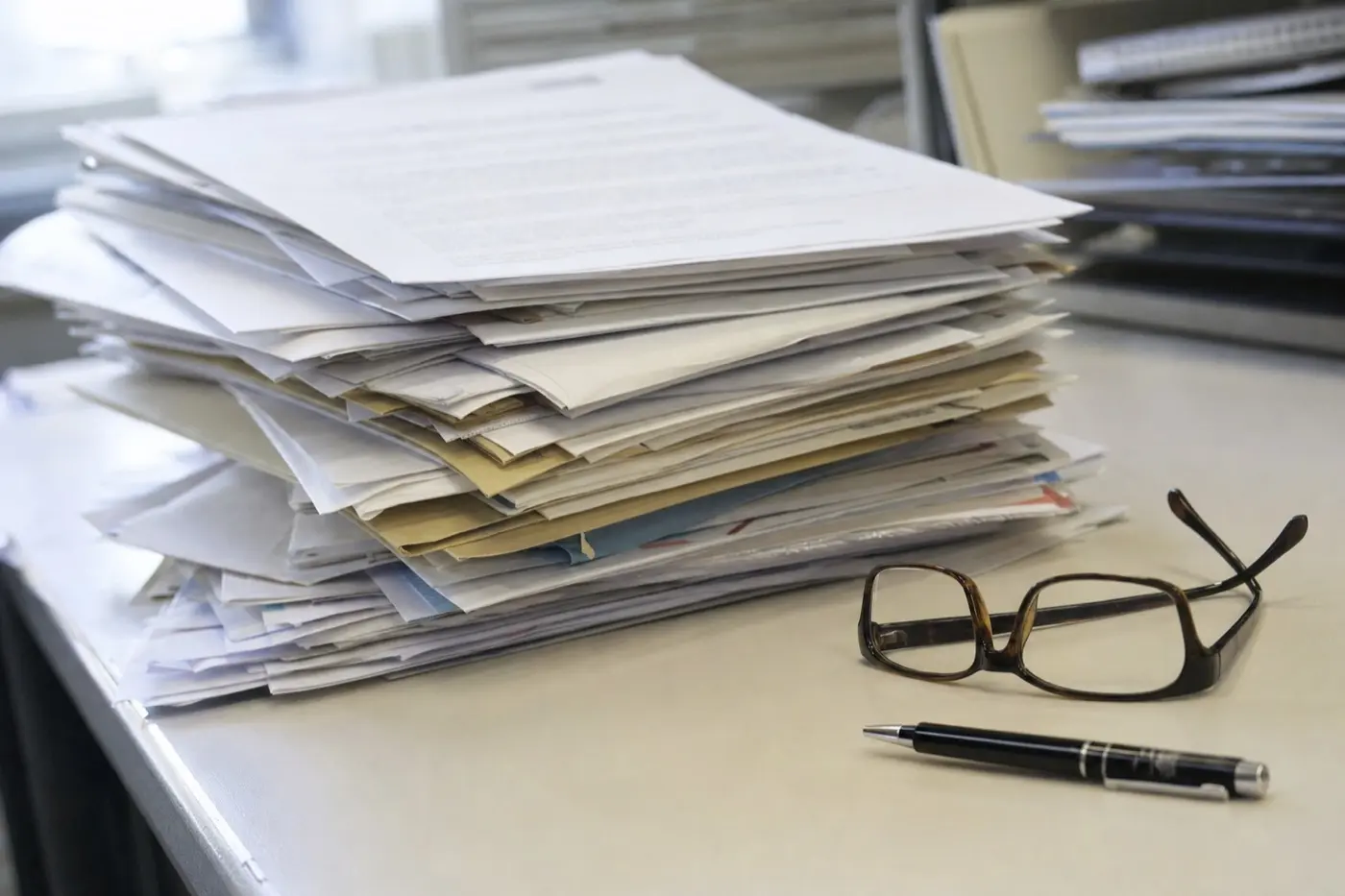 Stapel Briefe und ausgedruckter E-Mails auf einem Schreibtisch, daneben eine Lesebrille