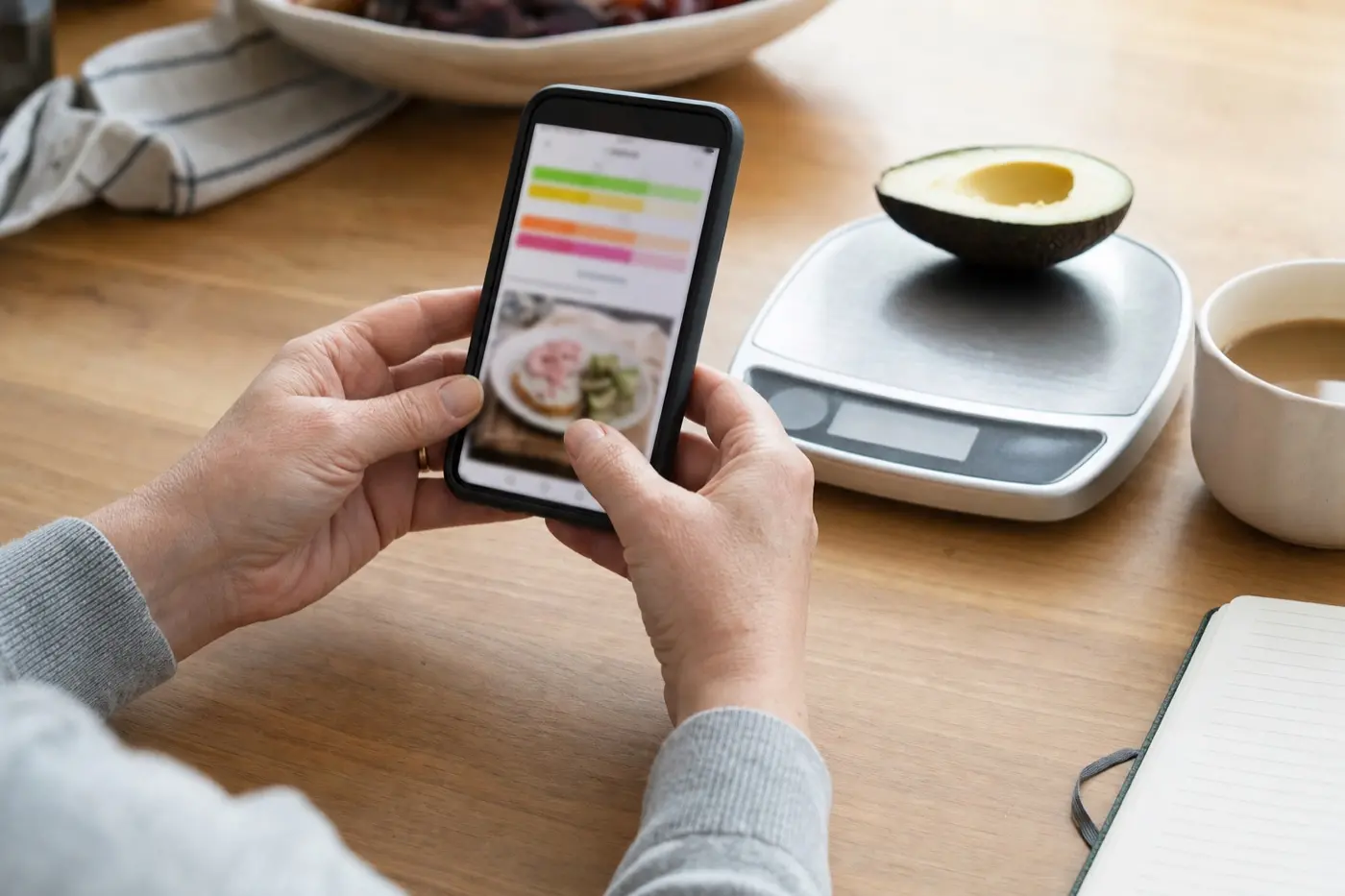 Hände einer Frau halten am Frühstückstisch ein Smartphone, daneben eine Küchenwaage mit einer halben Avocado.