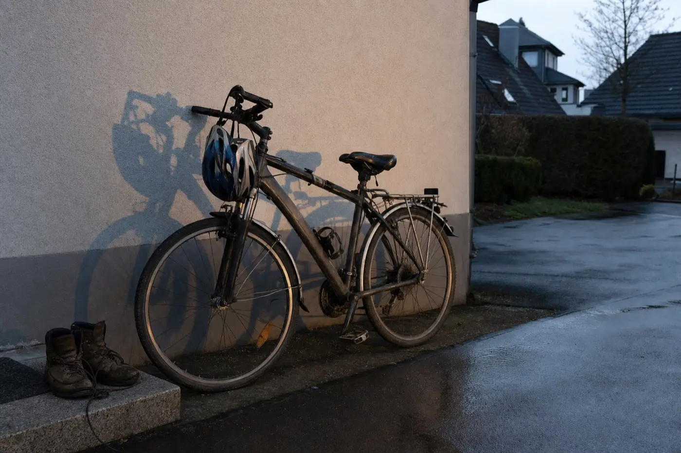 Herrenrad mit Helm am Lenker lehnt im Abendlicht an einer Garagenwand, daneben Arbeitsschuhe