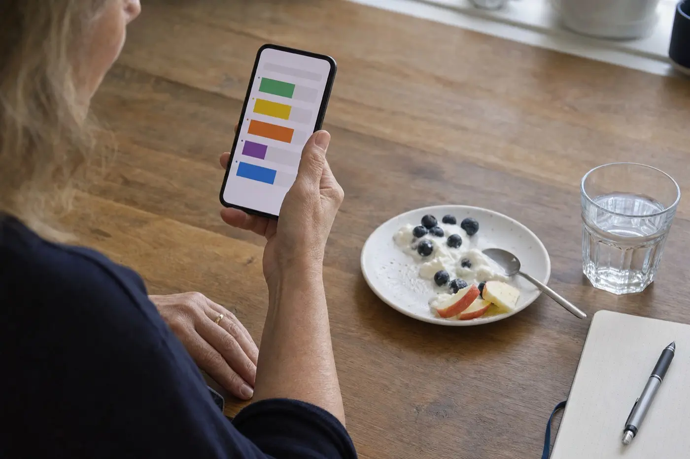 Küchentisch am Morgen: eine Frauenhand hält ein Smartphone mit einer Ernährungs-App, daneben ein kleiner Teller mit Quark und Obst und ein Glas Wasser.