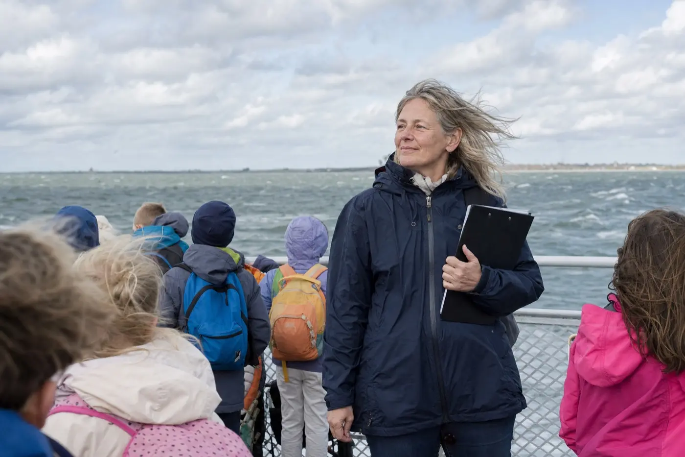Eine Frau um 50 in Regenjacke steht mit Grundschulkindern an der Reling einer Inselfähre.