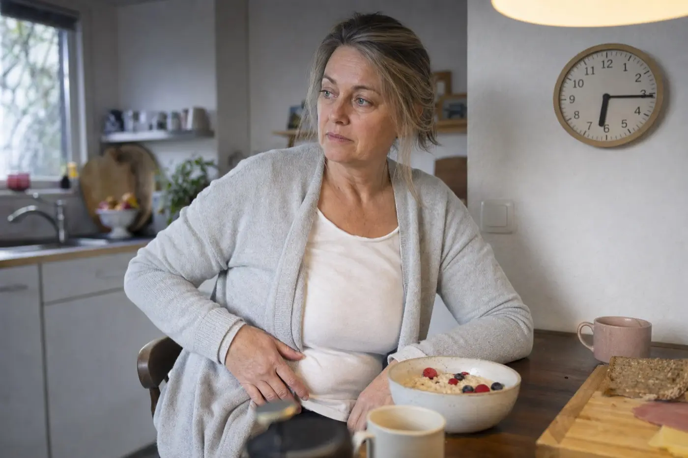 Frau um 50 am frühen Morgen in ihrer Küche, vor sich eine Schale Haferflocken mit Beeren, im Hintergrund eine Küchenuhr.