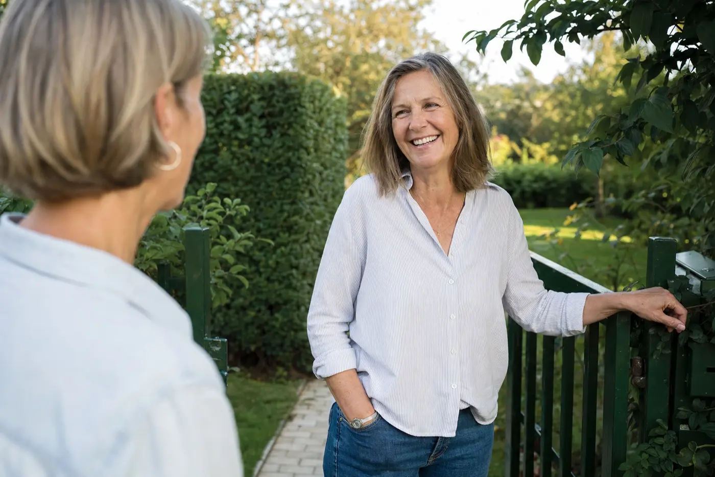 Frau Mitte 50 steht lachend an ihrer Gartenpforte im Gespräch mit einer zweiten Frau, die von hinten zu sehen ist.