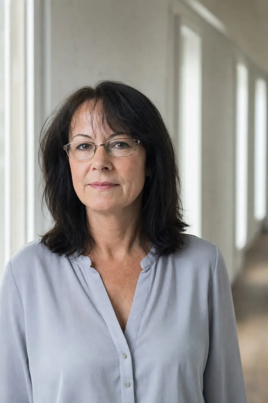 Porträt einer Frau um die 52 mit dunklem schulterlangem Haar und Brille in einem Büroflur.