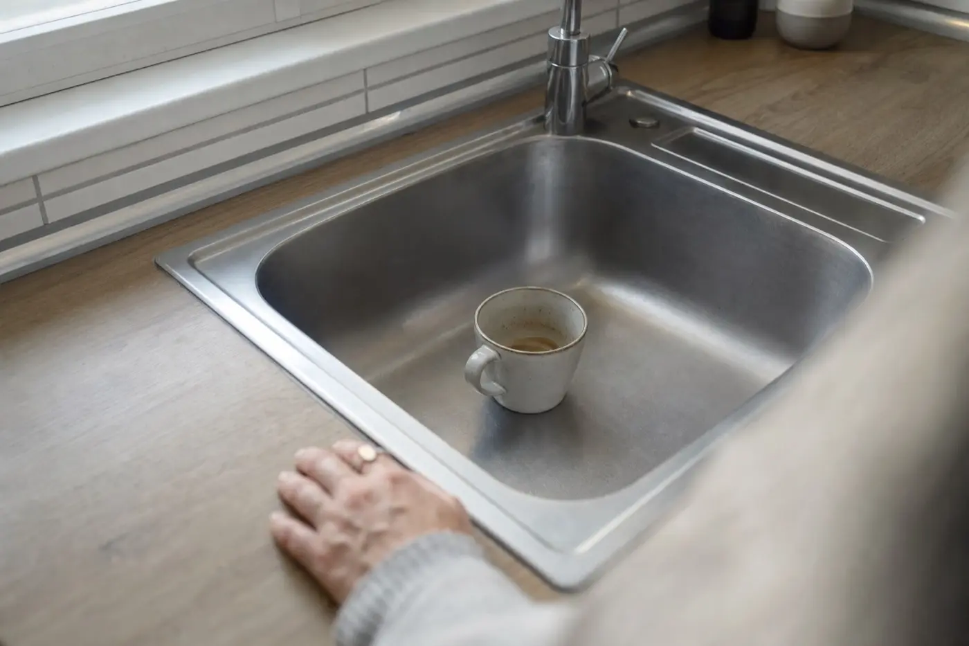 Einzelne Kaffeetasse in einer leeren Küchenspüle am Morgen, im Vordergrund unscharf eine Frauenhand auf der Arbeitsplatte